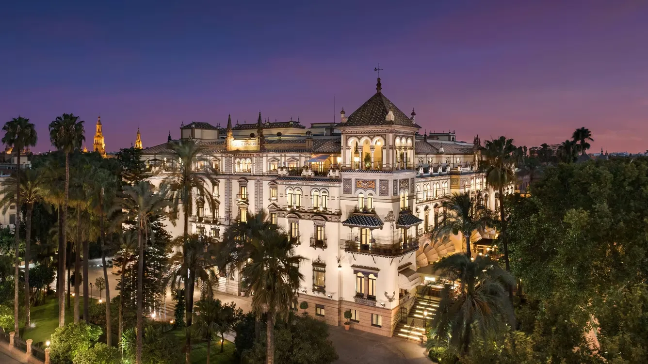Hotel Alfonso XIII at dusk, its tiled roofs and arcaded balconies lit above the palms of its garden, against a violet sky